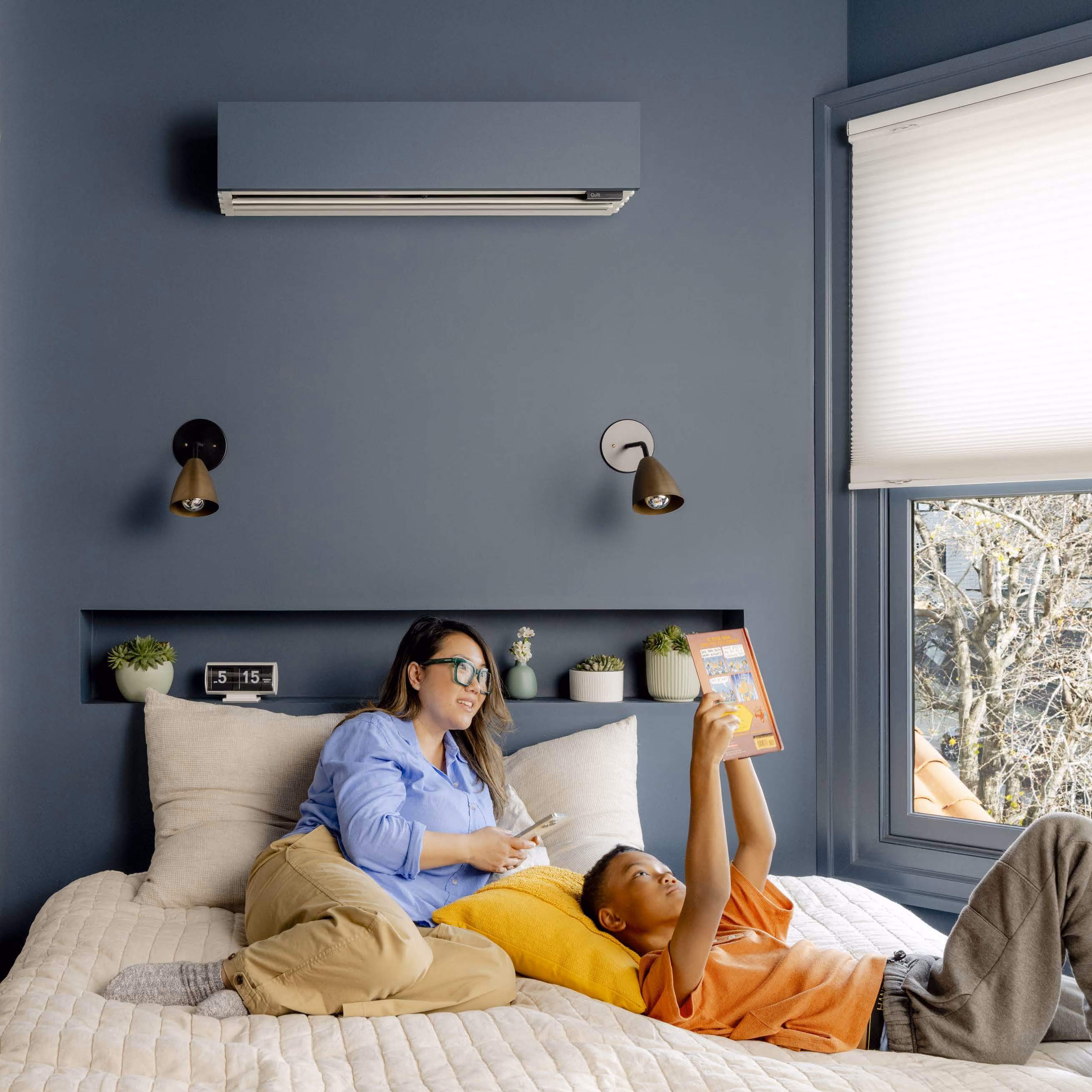 A woman in glasses and a blue shirt lies on her side on a beige quilted bed while a boy in an orange shirt lies next to her holding a book above him near a window with a view of trees.
