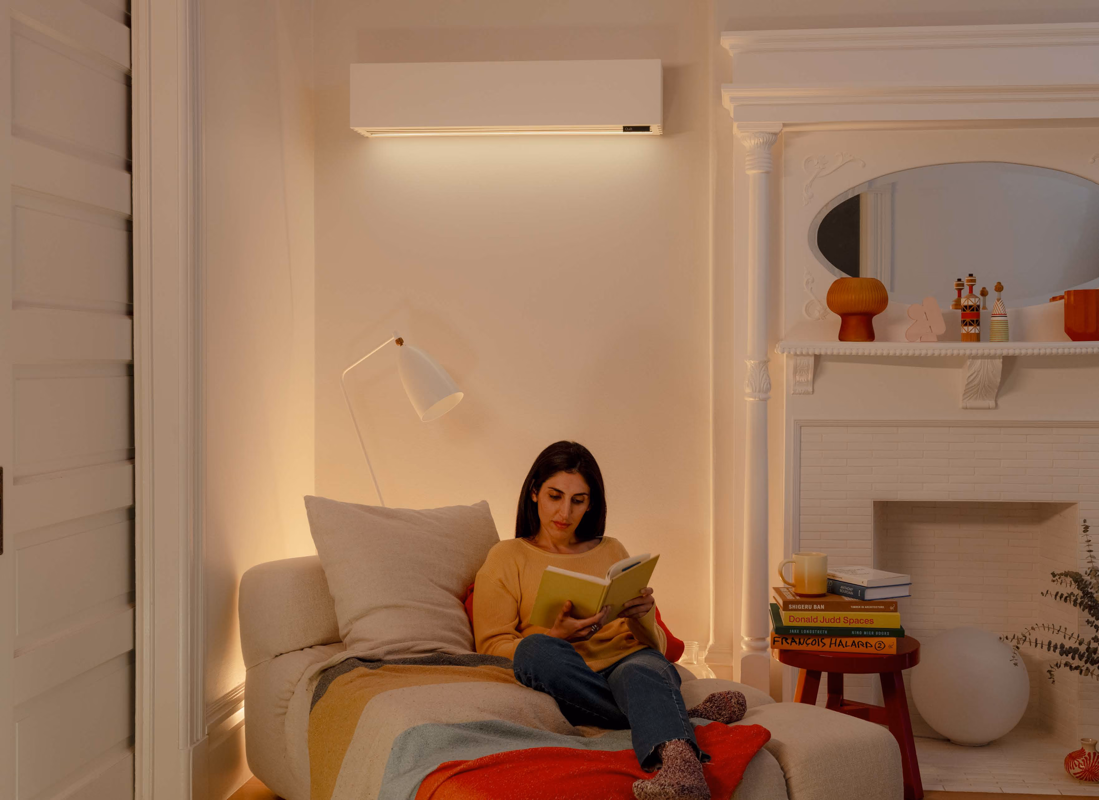 A woman with dark hair sits on a beige couch reading a book in a warmly lit living room with a white wall-mounted light above, a floor lamp behind her, and a white fireplace with decor and books stacked on a small wooden table.