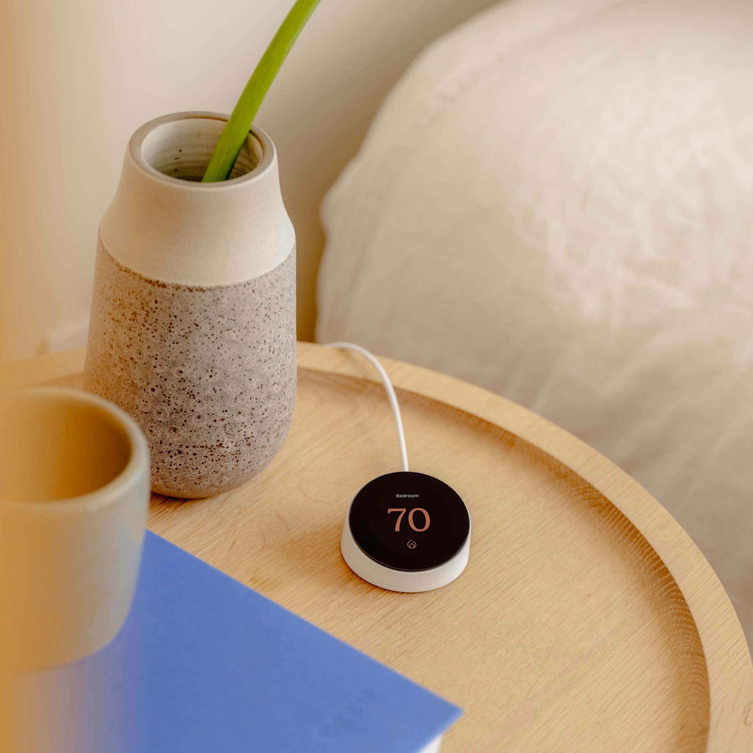 Round digital thermostat displaying 70 degrees on a wooden bedside table next to a grey textured vase with a green plant stem and a blue book, with a white bed in the background.
