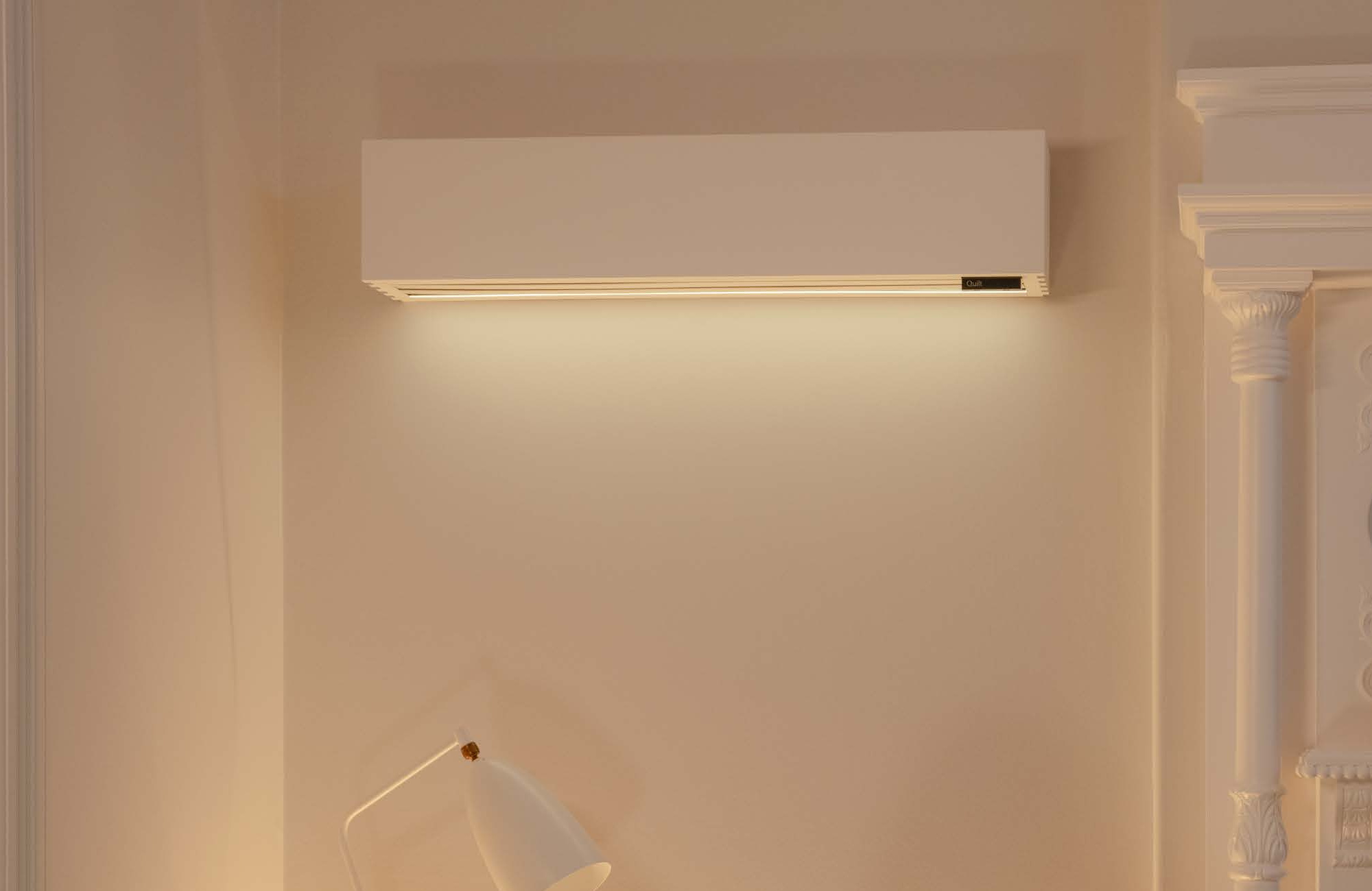 Modern rectangular wall-mounted light fixture emitting warm light onto beige wall, with a white lamp partially visible below and classic white decorative molding on the right.
