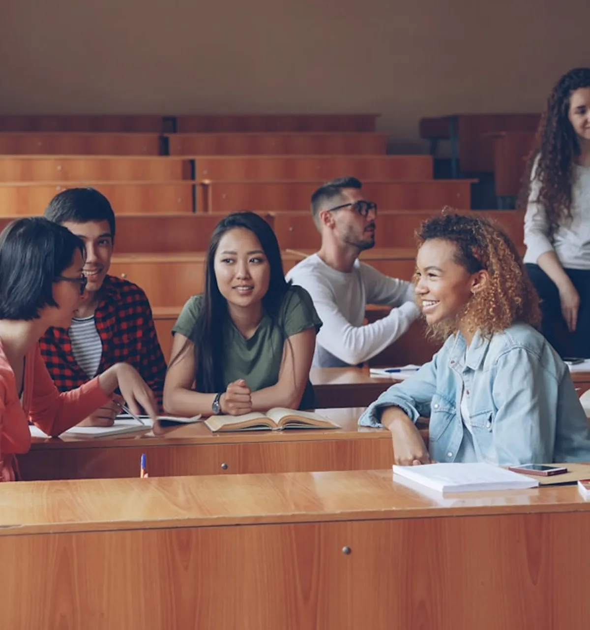 Group of diverse college students sitting in a lecture hall, talking and smiling with open books on desks.