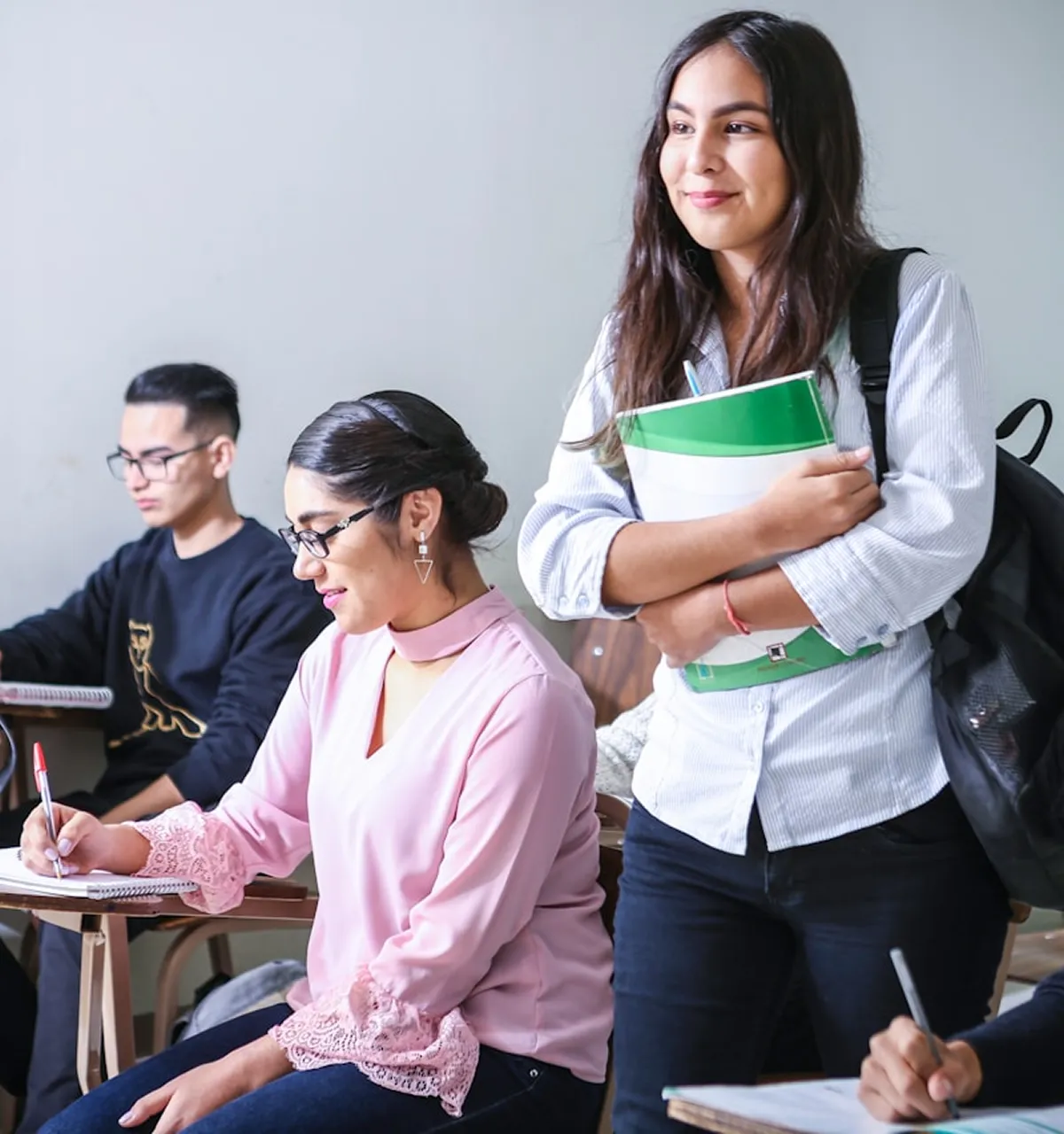 Students in a classroom, one standing and smiling holding notebooks, others sitting and writing at desks.