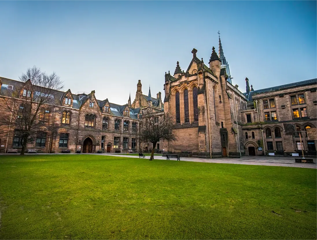 Historic stone university buildings with large arched windows surrounding a green lawn under a clear sky.