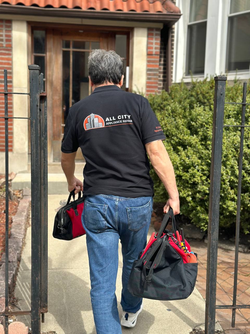 Person wearing a black 'All City Appliance Repair' shirt walking through an iron gate carrying two black and red tool bags.