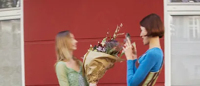 Two women smiling and exchanging a bouquet of flowers outdoors with a red wall background.