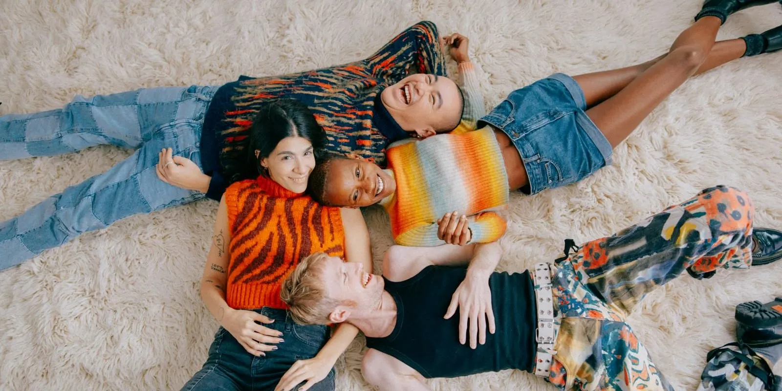 Four diverse young adults laughing and lying closely together on a fluffy white rug, wearing colorful casual clothing.