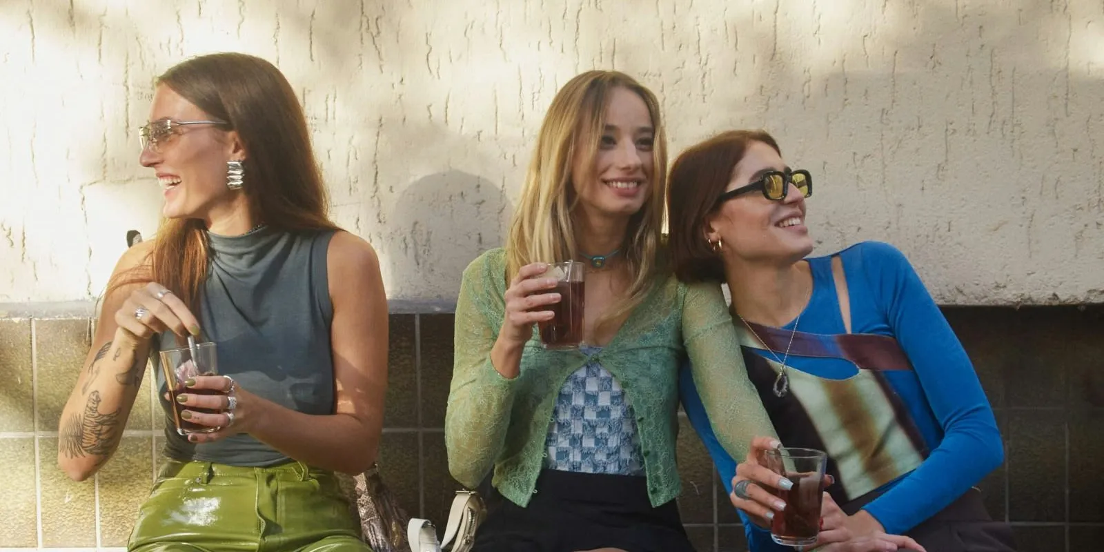Three young women sitting side by side outdoors, smiling and holding drinks in clear glasses.
