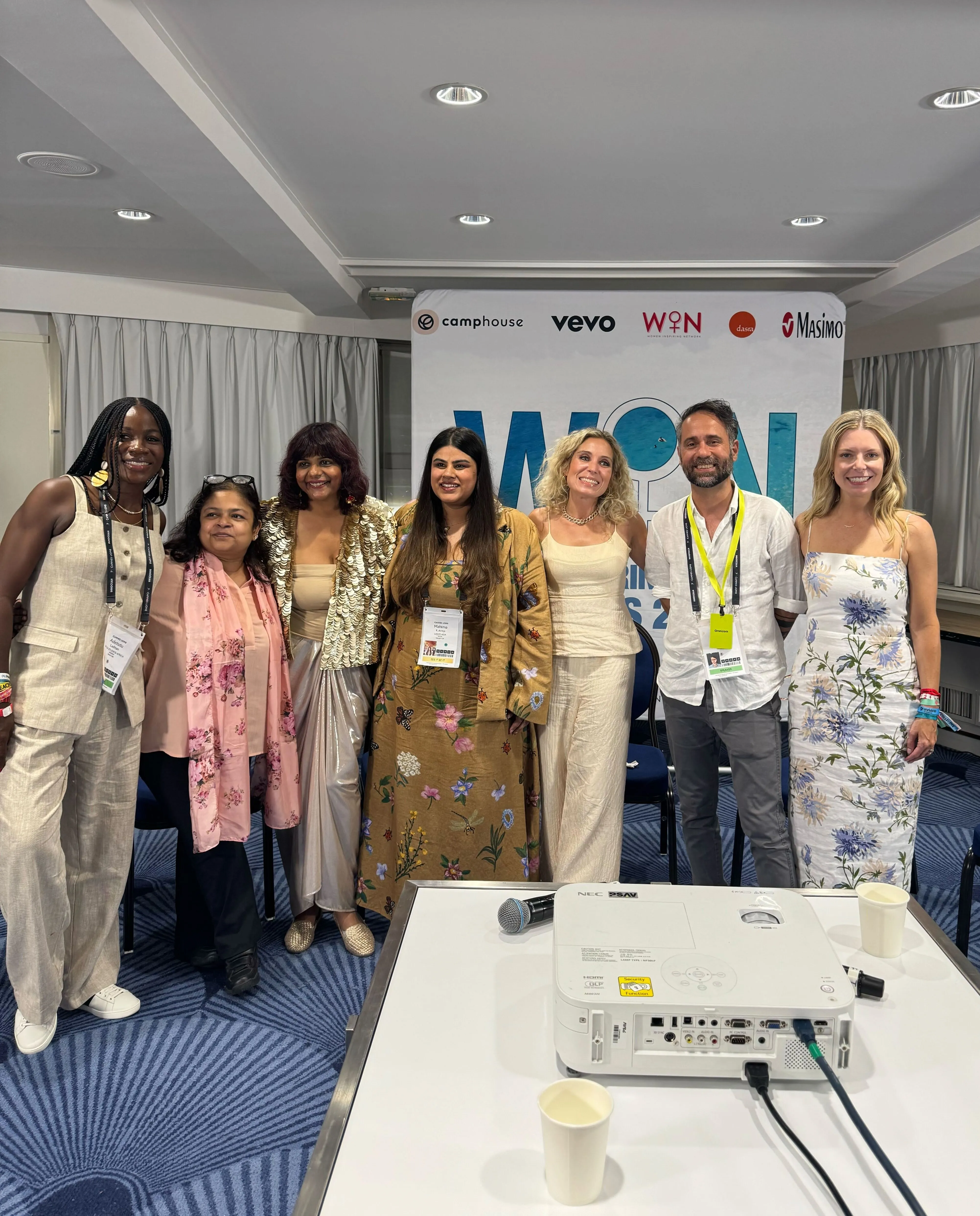 Seven people standing side by side in a conference room with a projector in the foreground and a banner with logos behind them.