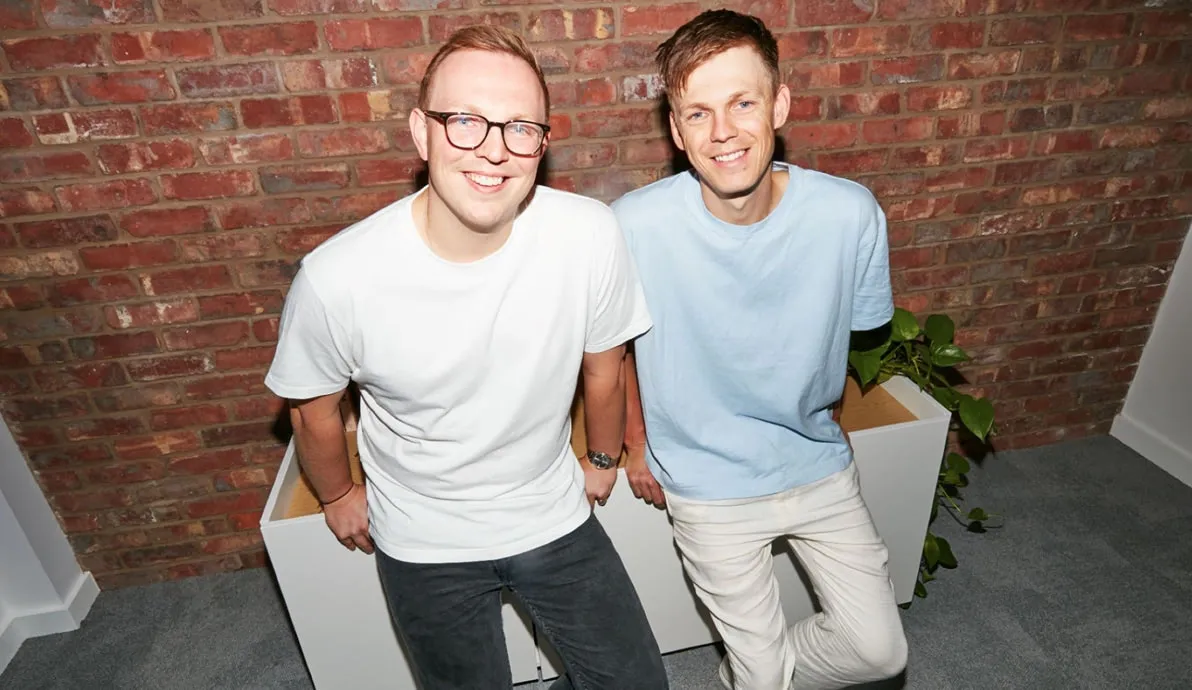 Two smiling men sitting on a white cabinet with a brick wall background indoors.