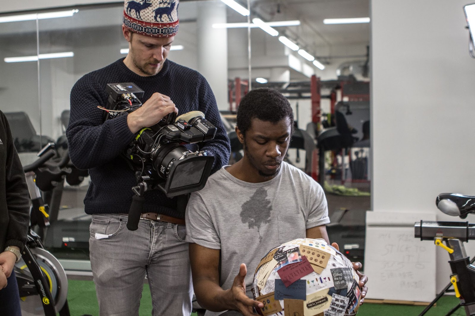 A freelancer filmmaker operates a camera while another person assists during a gym shoot, illustrating hands-on, compact video production.