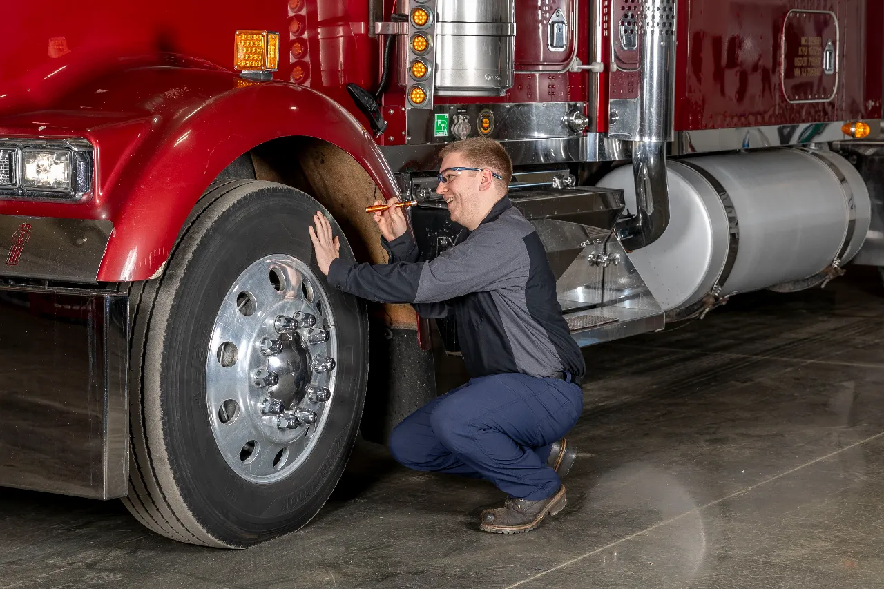 Mechanic performs DOT inspection on semi-truck, checking front tire tread depth with a gauge.