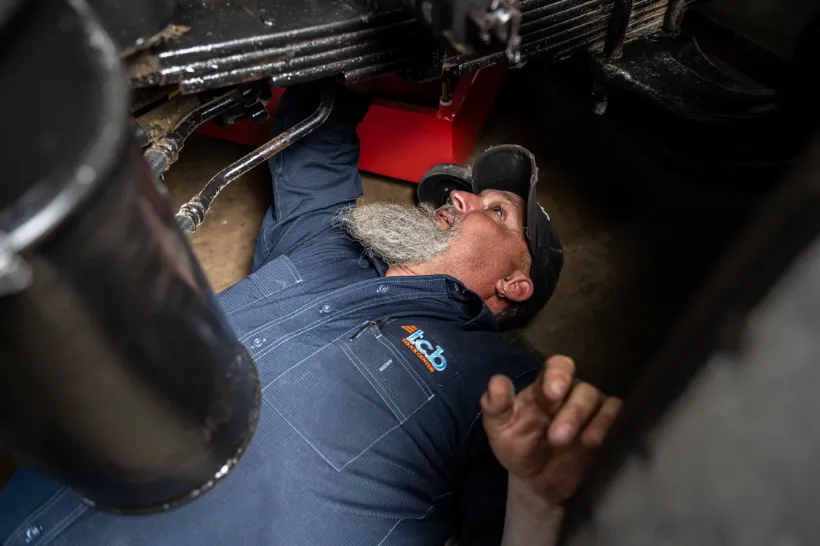 Technician performs DOT inspection under truck suspension, wearing a TCB-branded uniform shirt.