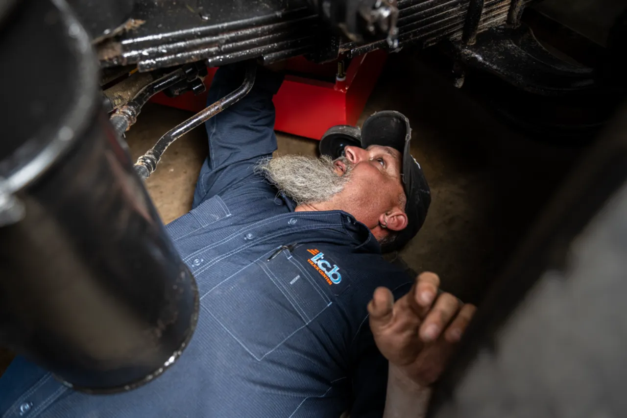 Technician performs DOT inspection under truck suspension, wearing a TCB-branded uniform shirt.