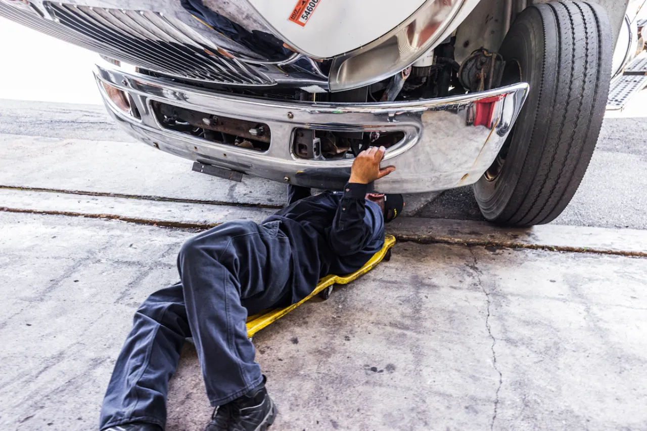 Technician performing preventive maintenance under a commercial truck using a creeper near the front bumper.