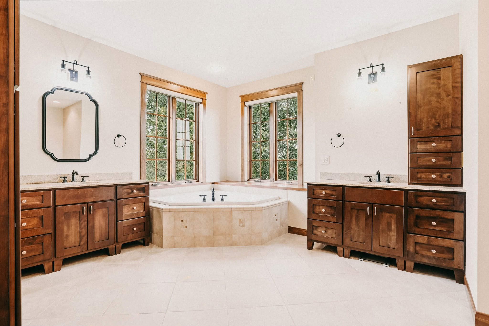 Bright bathroom with warm wood vanities, cream walls, and a corner soaking tub beneath two windows.