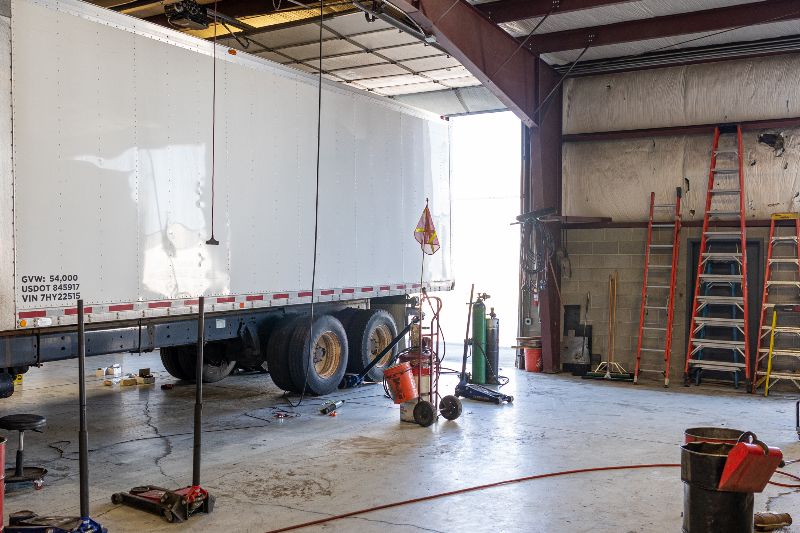 Interior of a truck repair shop with a white semi-trailer, ladders, tools, and welding equipment under natural lighting