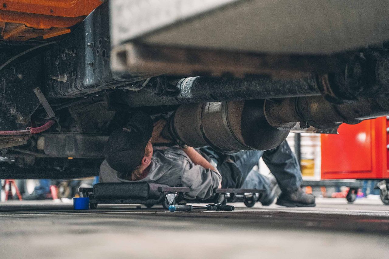 Mechanic lying on a creeper beneath a large truck inspects exhaust components and performs repair work in a busy diesel repair shop