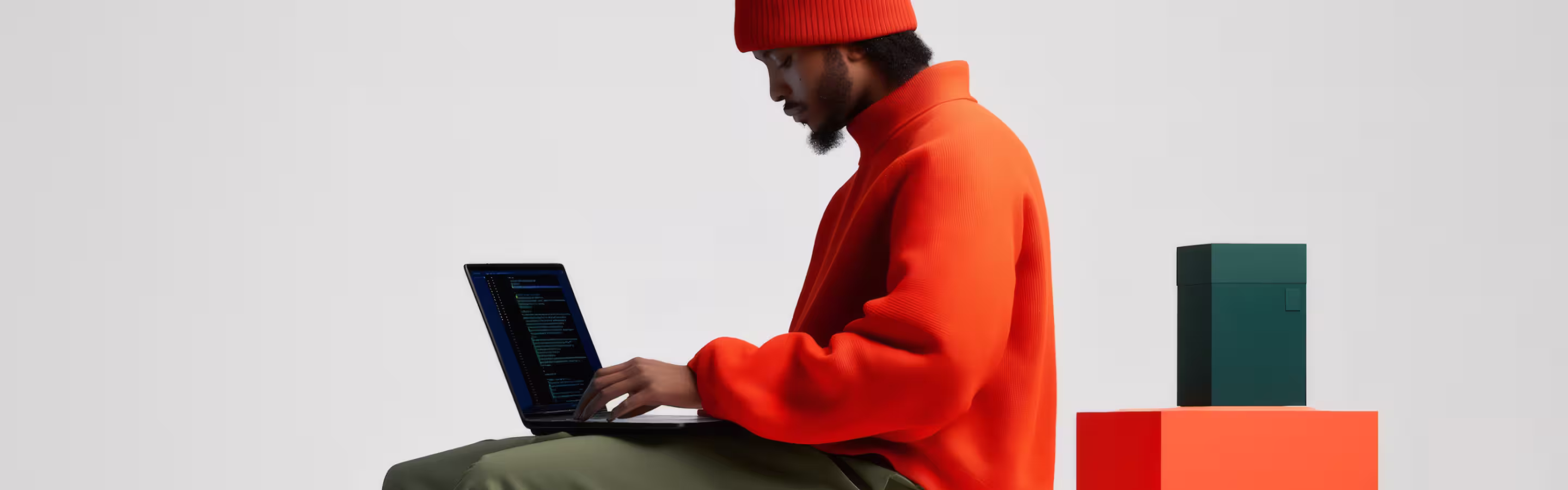 Side profile of a person wearing a bright red sweater and beanie, working on a laptop with code visible on the screen, sitting against a minimal white background with colorful blocks nearby.