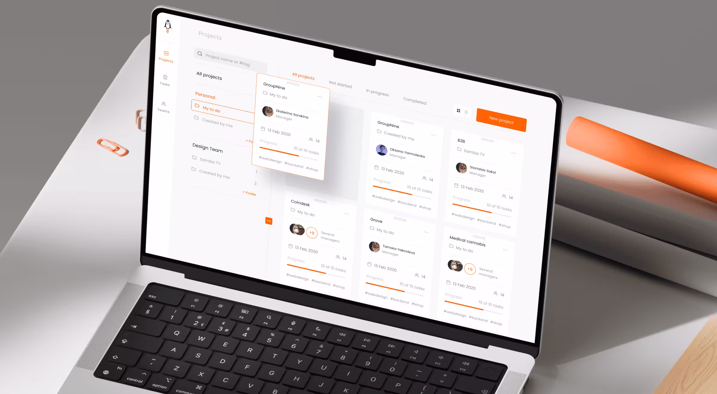 A sleek laptop screen displays a modern project management dashboard with a clean white and orange interface. The screen shows multiple project cards organized into columns under tabs like "All projects" and "In progress." Each card includes project names, managers, deadlines, progress bars, and tags such as #webdesign and #backend. The layout emphasizes clarity and task tracking, with a visible “New project” button on the top right. The background features a minimal desk setup with paper clips and orange design tools.