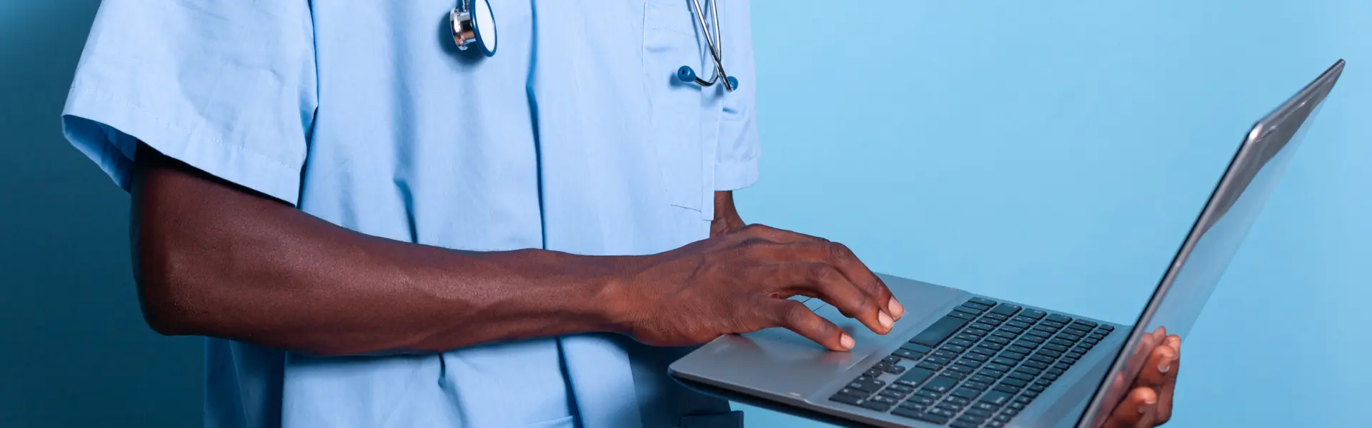 A healthcare professional wearing scrubs and a stethoscope types on a laptop against a blue background. The image suggests the use of digital tools in medical or clinical settings.