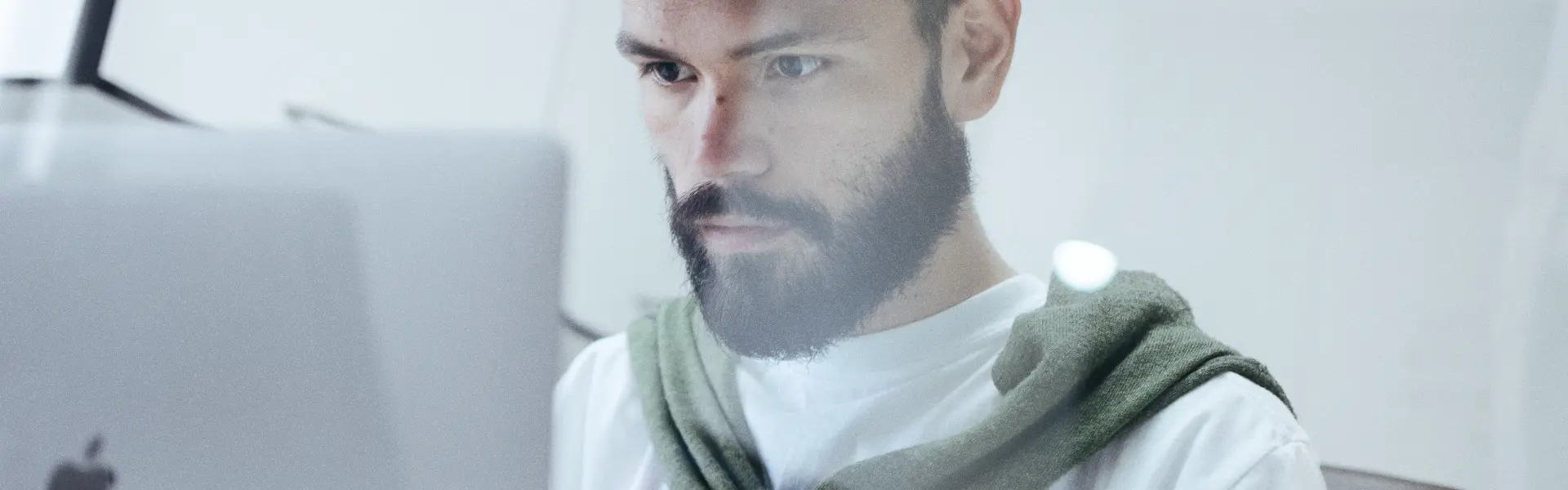 Man with a beard wearing a white shirt and green sweater draped over his shoulders, intently working on a silver Apple MacBook in a bright, modern workspace.
