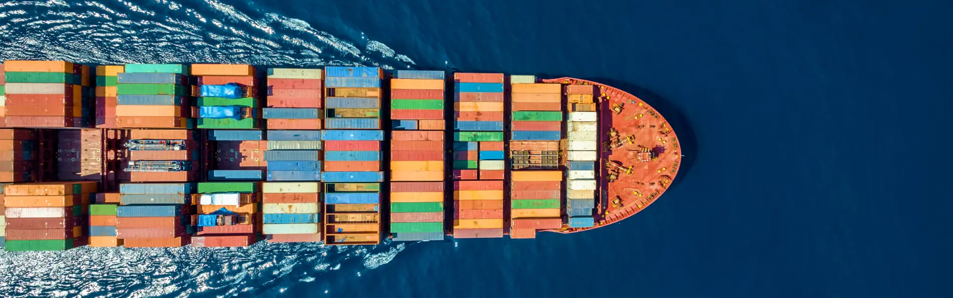 Aerial view of a large cargo ship traveling through deep blue ocean waters, carrying numerous colorful shipping containers neatly stacked on its deck. The ship is moving forward, leaving a white wake behind, indicating motion and direction. The vibrant colors of the containers contrast sharply with the surrounding sea, highlighting global trade and maritime logistics.