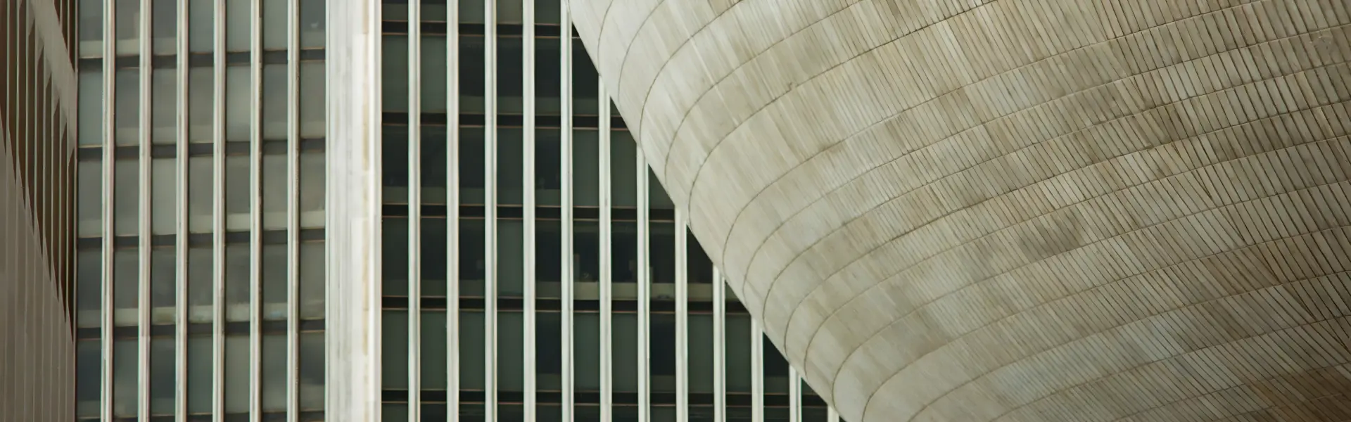 Architectural close-up showing the contrast between a modern high-rise building with vertical glass and steel window panels in the background, and a large, curved, ribbed concrete structure in the foreground. The juxtaposition highlights the interplay between linear geometry and sweeping organic form in urban architecture.