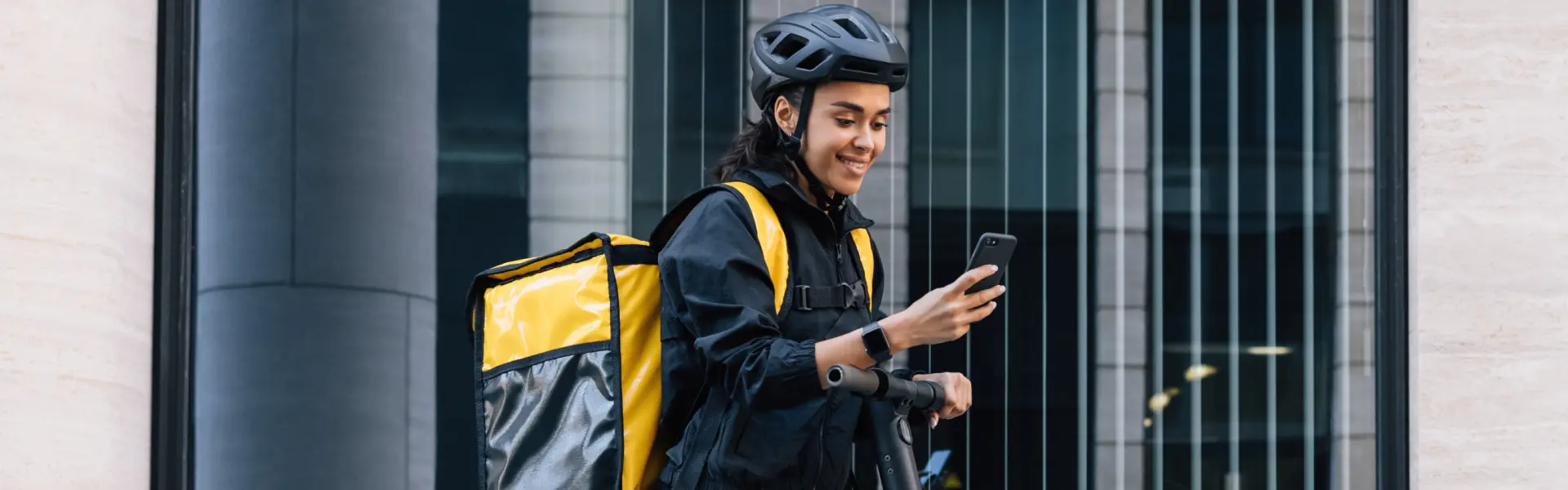 A smiling delivery person wearing a helmet and black-and-yellow uniform looks at their smartphone while holding the handlebars of an electric scooter. They are carrying a large yellow insulated backpack, likely used for food delivery, in an urban setting.
