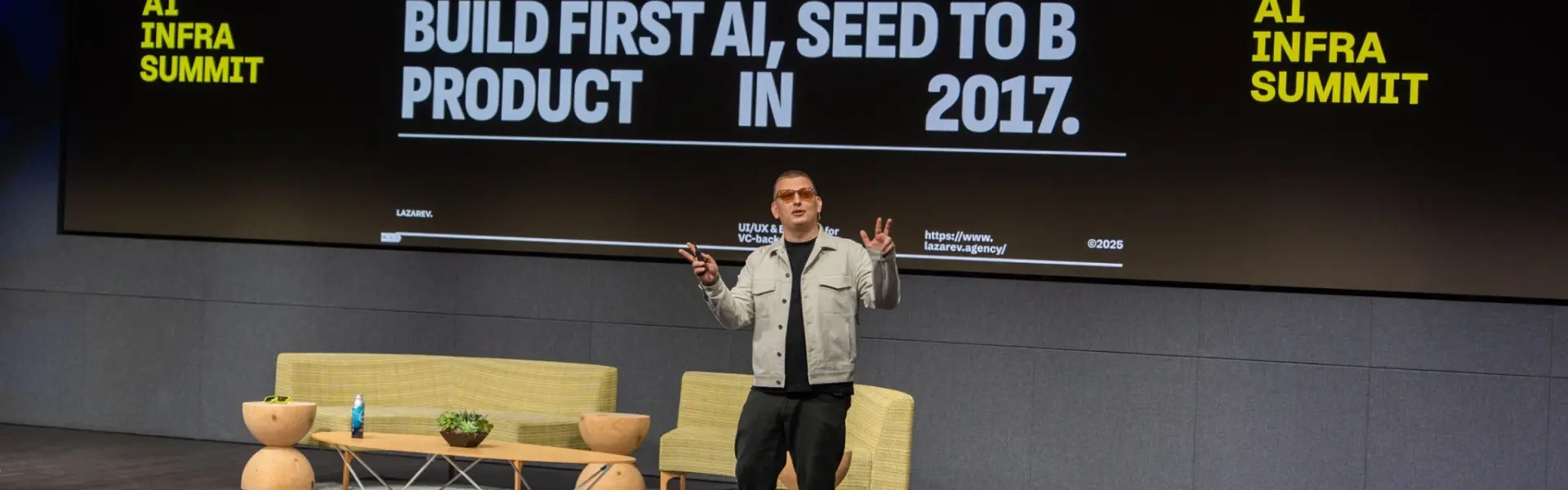 A speaker stands on stage at the AI Infra Summit, presenting in front of a large screen that reads: "100+ Awards Won. Build First AI, Seed to B Product in 2017." The backdrop includes modern furniture such as two green sofas, a coffee table with plants and water, and a dark-themed wall. The speaker wears sunglasses, a light jacket, black pants, and sneakers, gesturing with both hands mid-talk. The screen also displays the website “https://www.lazarev.agency/” and branding for LAZAREV. 