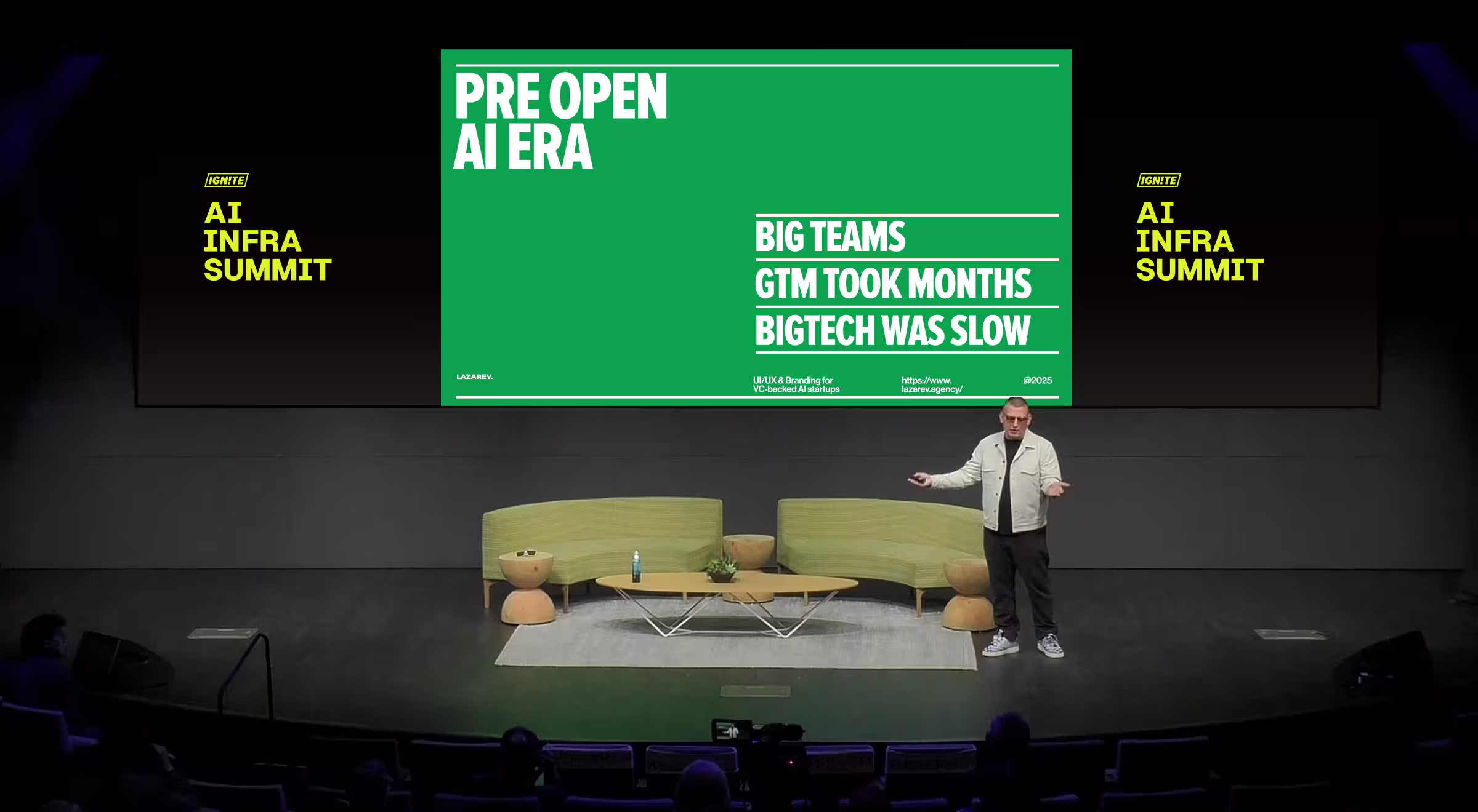 A speaker presents on stage at the AI Infra Summit, standing in front of a large screen with a green slide titled “Pre Open AI Era.” The slide highlights three key points: “Big Teams,” “GTM Took Months,” and “BigTech Was Slow.” The setting includes modern lounge furniture—two green sofas, wooden side tables, and a low coffee table with a water bottle and potted plant. The presenter wears a light jacket, black pants, and sneakers, gesturing with one hand. The screen also features the Lazarev agency branding and website: https://www.lazarev.agency/.