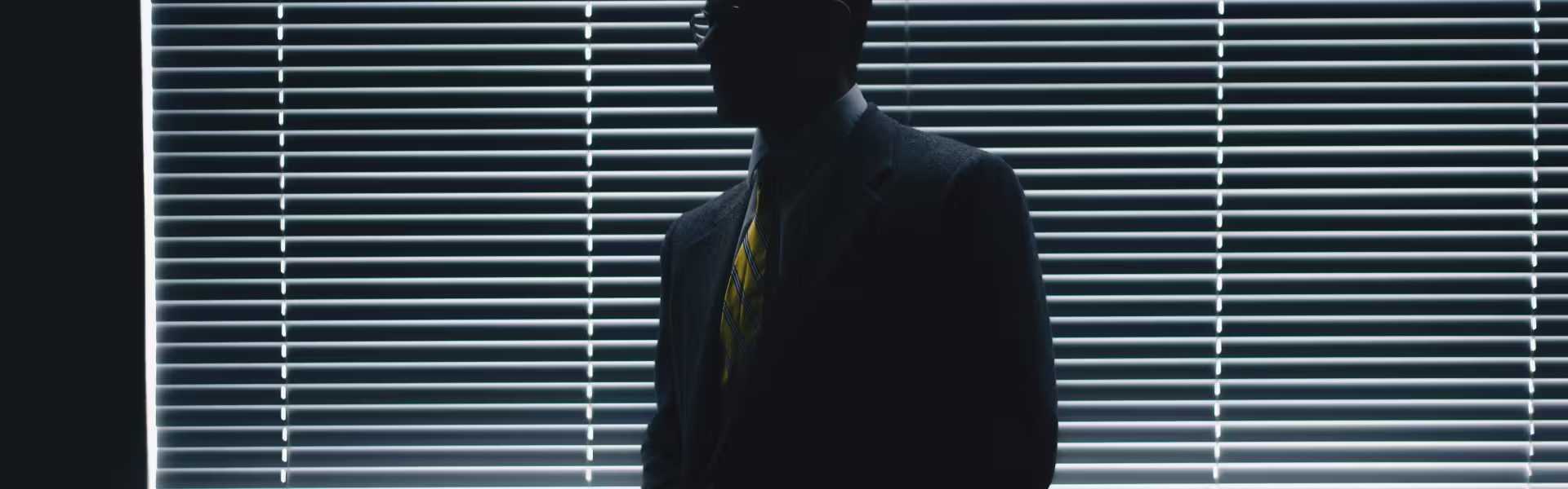 Silhouette of a man in a suit and striped yellow tie standing in front of closed window blinds, with light filtering through the slats creating a dramatic, shadowed effect.