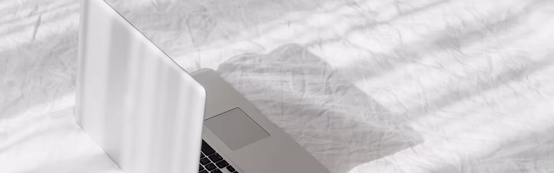 Open silver laptop on a white, sunlit bed with soft shadows cast by window blinds across the crumpled bedding.