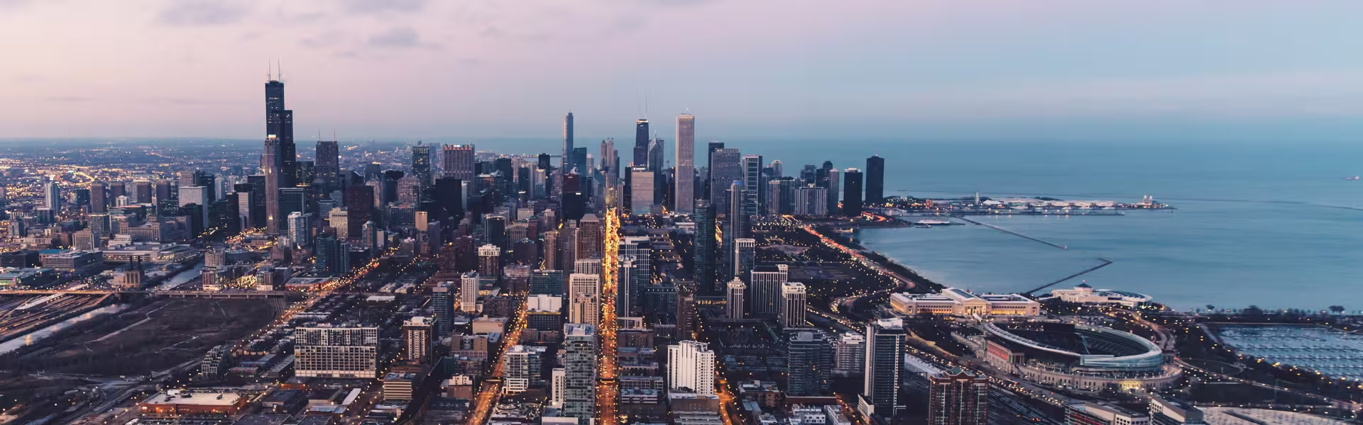 Aerial view of downtown Chicago at dusk, showcasing the city’s skyline with the Willis Tower and other high-rise buildings, illuminated streets, and Lake Michigan’s shoreline with Navy Pier and Soldier Field visible in the foreground.