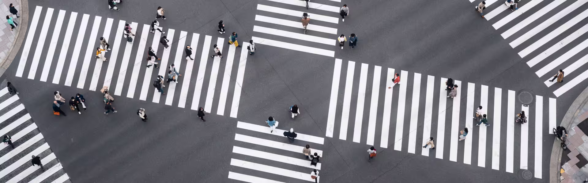 Aerial view of people walking across a large, multi-directional crosswalk with wide white stripes on dark asphalt, creating a geometric pattern at the intersection.