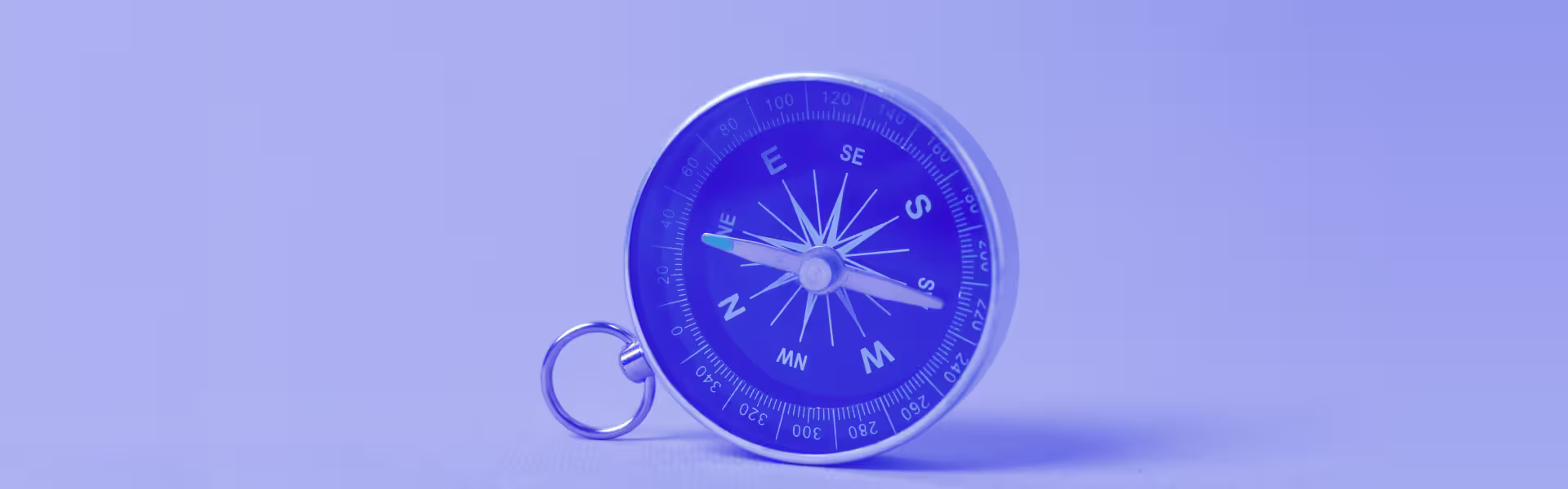 Close-up of a silver and blue compass with a keyring, placed on a flat surface, with its needle pointing northwest, set against a soft blue background.