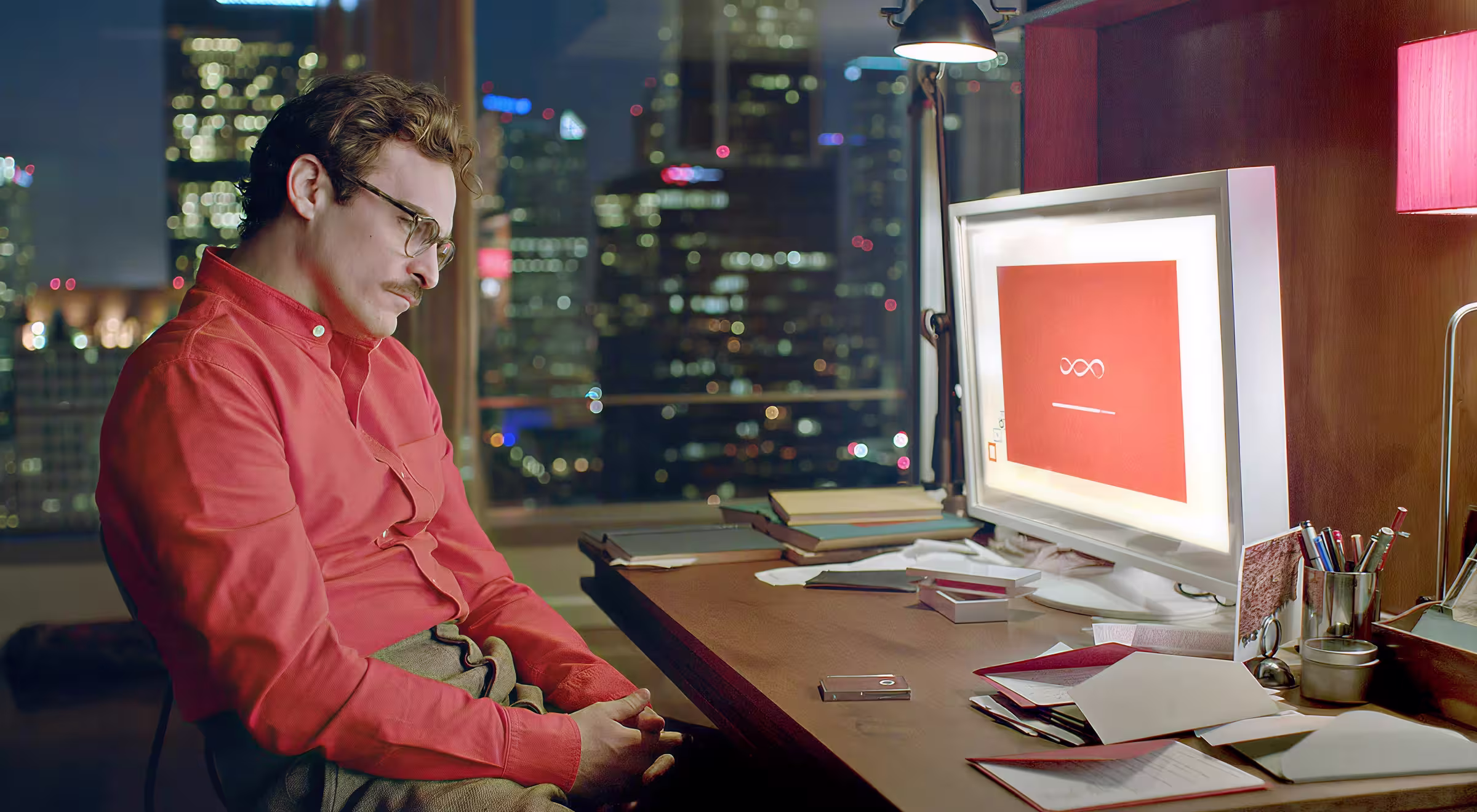 Man in red shirt sitting at desk at night, looking at computer screen with abstract AI loading symbol, city skyline in the background