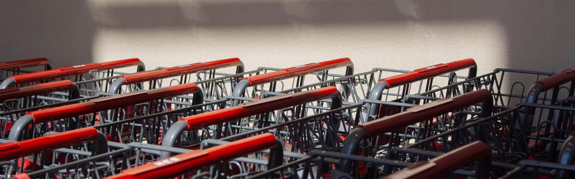 Row of red-handled shopping carts lined up outdoors in sunlight against a beige wall