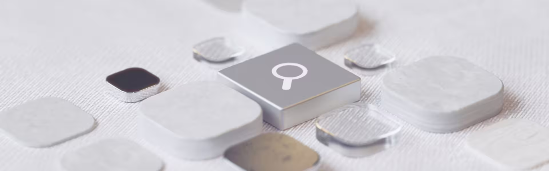Minimalist 3D interface elements on a light textured surface, centered on a raised square tile with a magnifying glass icon, surrounded by softly rounded translucent and metallic tiles suggesting search, discovery, or analysis