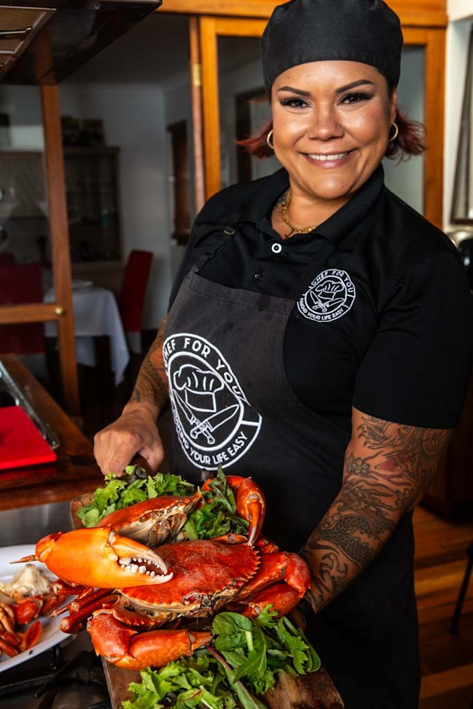 Smiling woman chef, Brita Brundin wearing he black Chef For You uniform holding a wooden board with a large cooked crab and fresh greens.