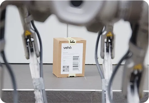 Cardboard package labeled 'Vehö' on a conveyor belt with robotic arms in the foreground.