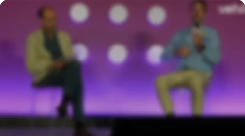 Two people seated on stage with a purple background, engaged in a conversation or interview.