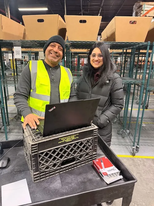 Two people standing in a warehouse with a laptop on a crate; one wears a yellow safety vest and the other a black jacket.