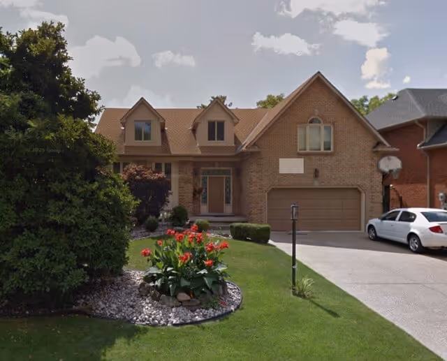 Two-story brick house with a double garage, manicured lawn, flower bed with red flowers, and a white car parked in the driveway.
