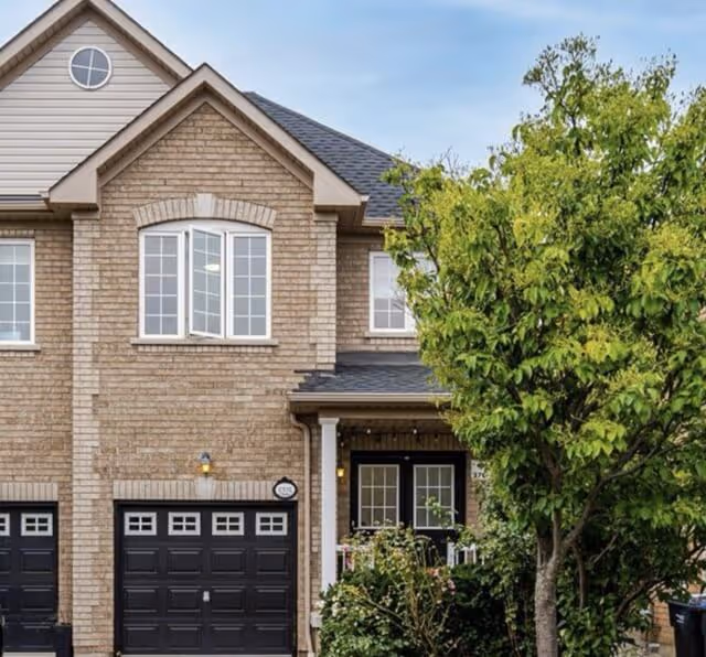 Two-story house with beige brick exterior, black garage doors, white-framed windows, and a leafy green tree in front.