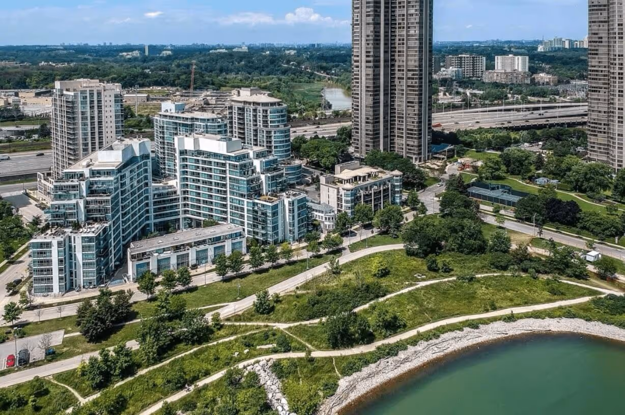 Aerial view of waterfront park with walking paths, green spaces, and modern high-rise residential buildings in the background under a clear sky.