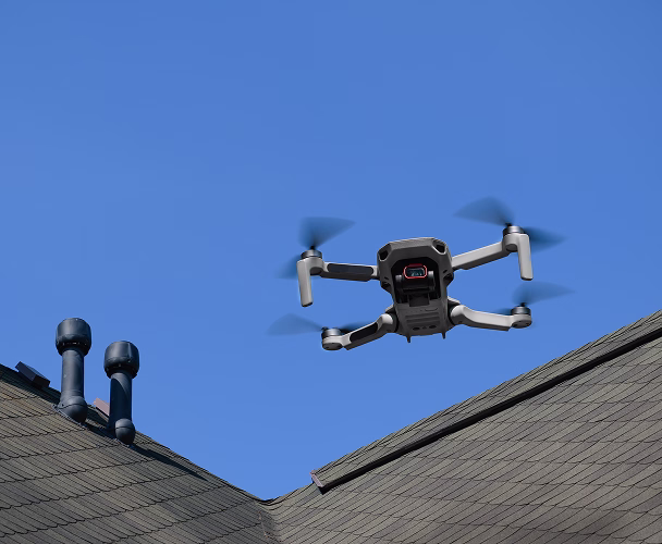 Small quadcopter drone flying above gray rooftops against a clear blue sky.