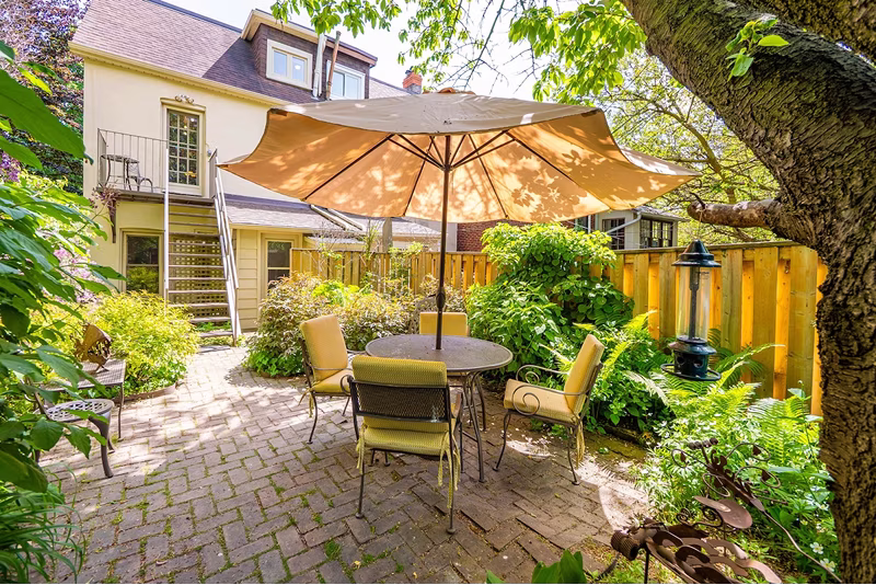 Cozy backyard patio with round table, four yellow cushioned chairs, large umbrella, and lush greenery.