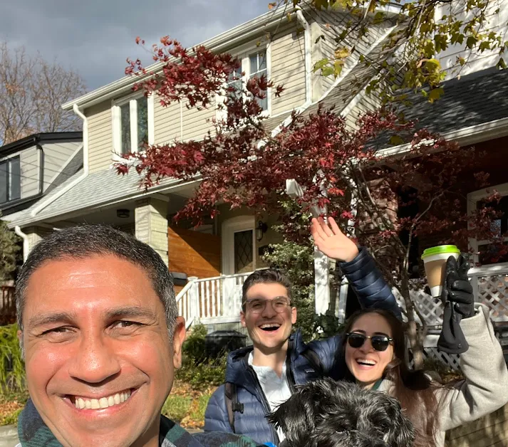 Three people smiling outdoors in front of a house with autumn trees, one holding a coffee cup and a dog.