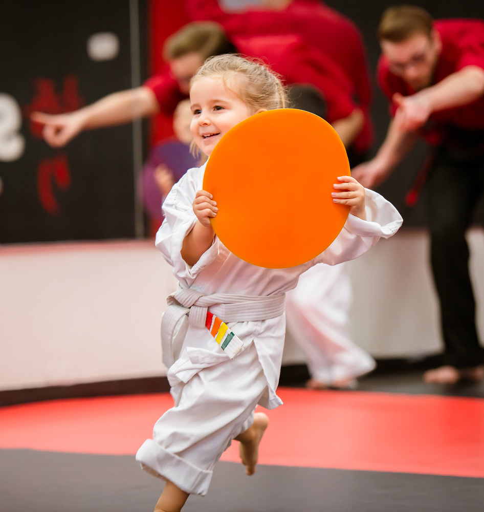 A Small child in karate gi smiling and running with an orange disc