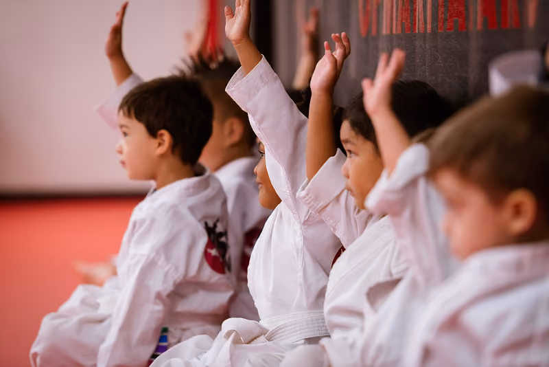 A row of children sitting in karate gi with their hands raised, asking a question
